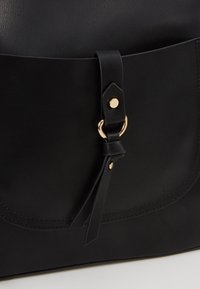 Czarna skórzana torba, zbliżenie na złoty pierścień i frędzle na klapie, z przeszyciami wzdłuż krawędzi.