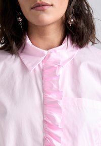 Light pink shirt featuring a pointed collar and a ruffled placket. Fabric appears smooth with a subtle sheen. Pocket on the left side.