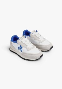 Zapatillas blancas con acentos azules, presentando una puntera de ante gris texturizado, un forro interior azul y suelas de goma negras. Logo de calavera en el lado.