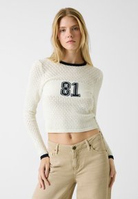 Gebreide pullover trui in crème met een zwarte geribbelde halslijn en een cropped ontwerp, met het nummer "81" in marineblauw over de borst.