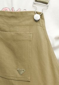 Μπεζ υφασμάτινη φόρμα με τσέπη στο μπροστινό μέρος και κεντημένο λογότυπο καρδιάς, κλειστή με ασημένιο μεταλλικό κούμπωμα σε λευκό φόντο.