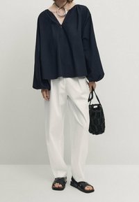 Marineblått löst sittande blus med v-ringning, vita vida byxor, svarta platta sandaler och en texturerad svart handväska. Enkla designinslag noteras.