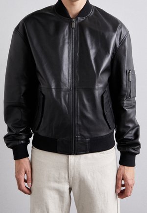 Mann trägt eine schwarze Leder-Bomberjacke mit Frontreißverschluss, Seitentaschen und Reißverschlusstasche am Ärmel, kombiniert mit hellbeigen Hosen.