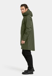 Olivgrön vattentät parka med huva, lång silhuett, sidofickor och elastiska muddars. Bärs över svarta byxor och stövlar.