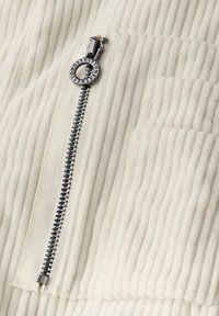 Tissu en velours côtelé blanc avec des côtes verticales, comportant une fermeture éclair en métal noir avec un tirette circulaire et un logo sur la fermeture.