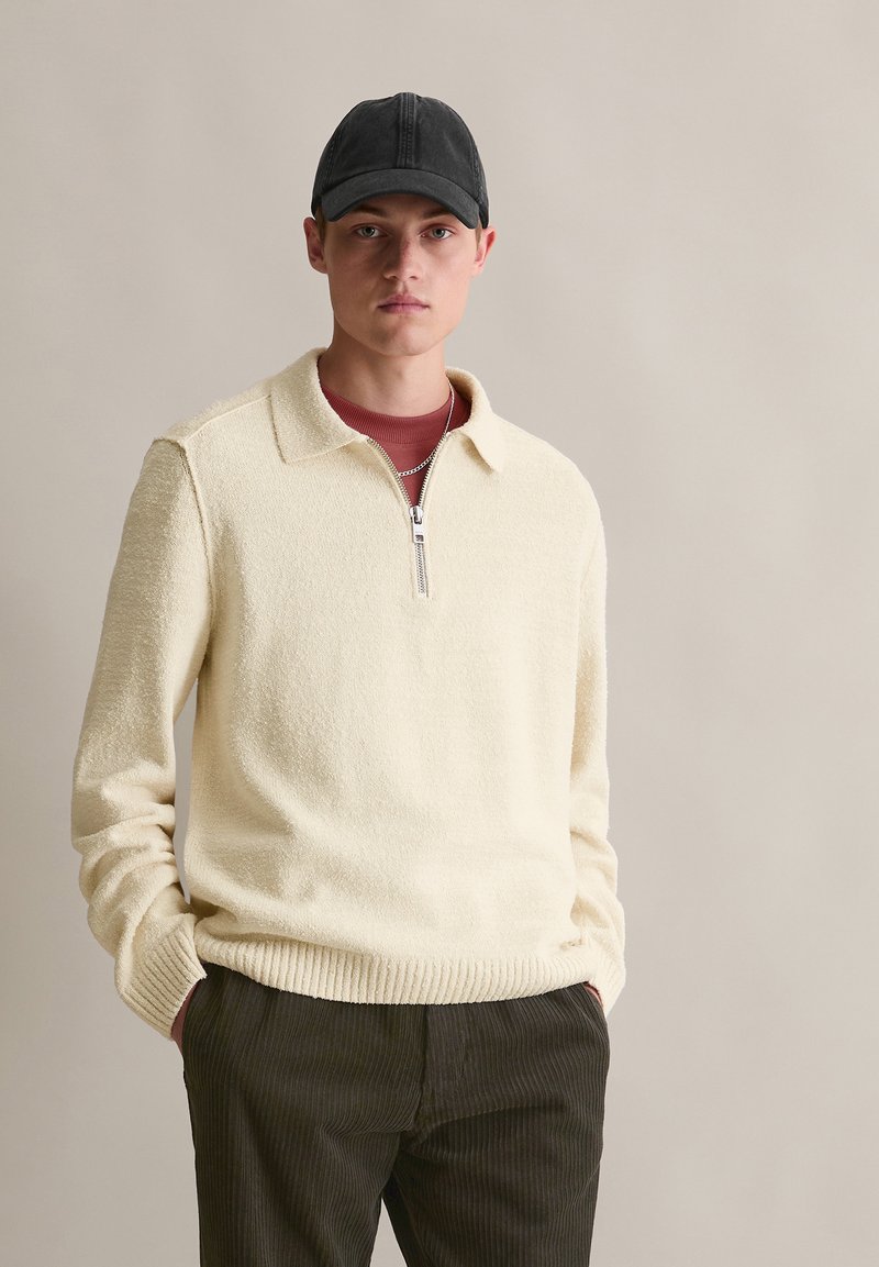 Sweter polarowy w kolorze kremowym z kołnierzem i półzipem, noszony z wiśniową koszulą pod spodem. Mężczyzna ma na sobie czapkę w kolorze czarnym oraz ciemne spodnie z kordurowej tkaniny.