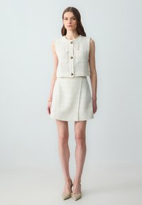 Witte cropped top met gouden knopen en twee voorzakken, gecombineerd met een bijpassende mini rok van textuur. Model draagt beige puntige hakken.