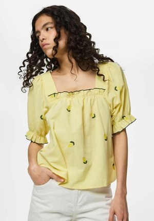 Femme aux cheveux bouclés et foncés portant un chemisier jaune à manches bouffantes avec une broderie de citrons, associée à un pantalon blanc, posant avec une main dans la poche.