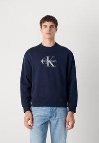 Sweatshirt azul-marinho feito de um tecido suave, apresentando um logótipo branco proeminente da Calvin Klein na frente. Gola redonda clássica e punhos canelados.