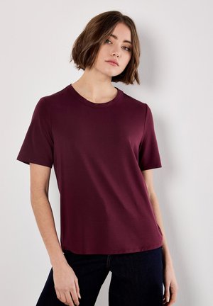 Mujer joven con el cabello corto y castaño, vistiendo una camiseta corta burdeos lisa y pantalones oscuros, apoyada contra una pared blanca.
