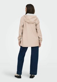 Chaqueta con capucha color beige claro, con cordones, puños elásticos y un corte holgado, combinada con jeans de pierna ancha azul oscuro y calzado beige.
