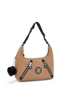 Tan-värinen olkalaukku mustilla yksityiskohdilla, jossa on hopeiset vetoketjut, pyöreä logo-patch ja musta pom-pom-avainketju.