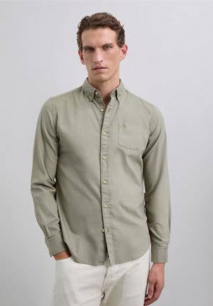 Hombre con cabello rizado castaño claro, vistiendo una camisa de botones verde claro y pantalones blancos, de pie contra un fondo gris claro liso.