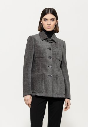 Frau in einem grauen, geknöpften Wollblazer mit vier Taschen über einem schwarzen Rollkragenpullover und schwarzen Hosen, die vor einem schlichten weißen Hintergrund steht.