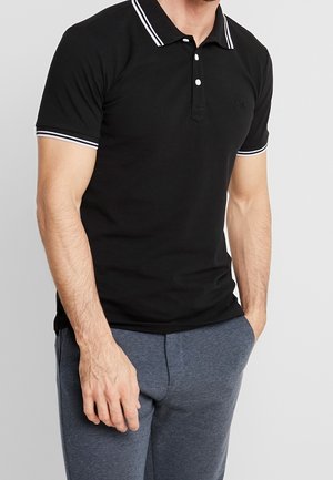Sort poloshirt med en tre-knaps lukning, hvide stribede detaljer på kraven og ærmerne, båret med grå joggingbukser. Blødt bomuldsstof.