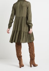 Olive green, długa sukienka z długim rękawem, o warstwowym dołku i luźnym kroju. Noszona z brązowymi zamszowymi botkami za kolano, z blokowym obcasem.