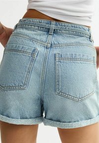 Los pantalones cortos de mezclilla azul claro presentan una cintura alta, dobladillos enrollados y dos bolsillos traseros con un sutil detalle de costura. Están hechos de un material suave y texturizado.