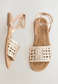 Flache Sandalen mit einem gewebten metallischen Riemen und einer weichen Beigen Suede-Innensohle. Verfügen über einen verstellbaren Knöchelriemen und einen niedrigen Absatz.