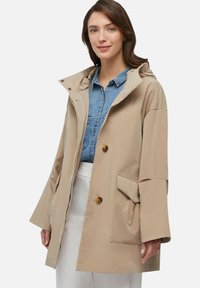 Impermeabile beige con cappuccio, dotato di maniche ampie, bottoni e una tasca frontale, realizzato in tessuto liscio. Abbinato a una camicia di denim sotto.