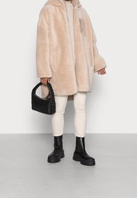 Beige faux pelsjakke med hætte og sidelommer, båret over lys beige top og cremefarvede leggings, kombineret med sort flettet håndtaske og kraftige sorte støvler.