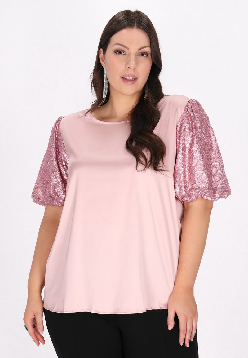 Lys pink bluse med korte, puffede, pailletbesatte ærmer, glat tekstur, rund udskæring og løs pasform. Sorte bukser synlige forneden.