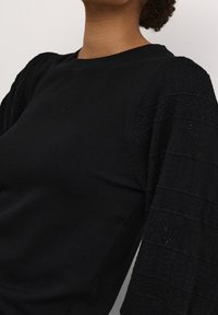 Svart långärmad topp med texturerade stickade ärmar med mönstrade detaljer och rundad halsringning. Tillverkad av ett mjukt, stretchigt material.