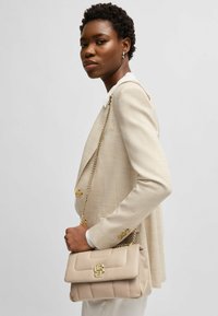 Borsa a tracolla in pelle beige con logo dorato, texture trapuntata e tracolla a catena, tenuta da una persona in un blazer di colore chiaro.