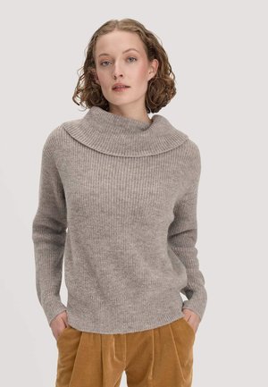 Jeune personne aux cheveux courts et bouclés portant un pull côtelé gris clair et un pantalon en velours côtelé couleur moutarde, les mains dans les poches.
