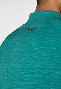 Αθλητική μπλούζα σε τιρκουάζ με λεπτό υφασμένο μοτίβο, που διαθέτει όρθιο γιακά και μαύρο λογότυπο στη ράχη του γιακά.