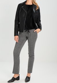 Schwarze Lederbikerjacke über einem schwarzen gefältelten Top, kombiniert mit grauen strukturierten Hosen und schwarzen Loafers. Model steht vor einem neutralen Hintergrund.