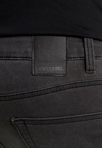 Svart denimjeans med en texturerad läderetikett med präglad branding. Sömnadsdetaljer är synliga och förbättrar den övergripande designen.