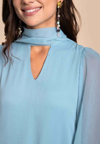 Lichtblauwe blouse met een keyhole-halslijn, gemaakt van zachte, gestructureerde stof en lange mouwen. Vergezeld van lange, sierlijke oorbellen.