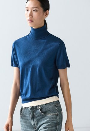 Femme portant un pull à col roulé bleu à manches courtes sur une chemise blanche et un jean en denim bleu clair sur un fond clair uni.