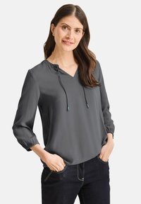 Blusa gris, ligera, con escote en V y cordón ajustable. Mangas tres cuartos con puños elásticos. Textura suave, combinada con jeans oscuros.