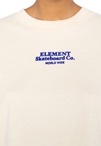Crème-kleurig katoenen T-shirt met blauwe geborduurde tekst: "ELEMENT Skateboard Co. WORLD WIDE" gecentreerd op de borst.
