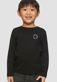 Lachende jonge jongen in een zwart T-shirt met lange mouwen en witte ronde tekst op de borst, met donkere spijkerbroek, staand tegen een effen achtergrond.