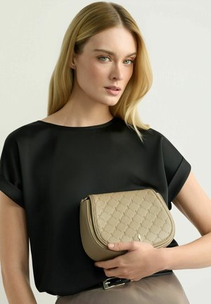 Kobieta o długich blond włosach, ubrana w czarną bluzkę, trzymająca beżową pikowaną torebkę na jasnym, jednolitym tle.