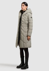 Padded long coat in light gray with a large hood, quilted texture, diagonal seams, and black accents. Worn over a black top and tights.