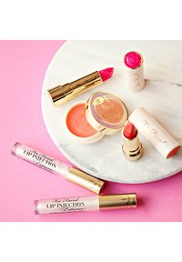 Too Faced lipproducten, waaronder een roze lippenstift, een perziken lip scrub en glans, gepresenteerd op een marmeren oppervlak met gouden accenten.