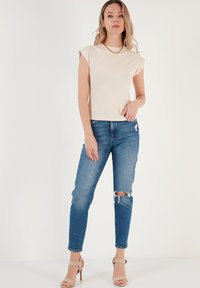 Top beige a maniche corte abbinato a jeans blu aderenti, con dettagli strappati sulle ginocchia. La modella indossa sandali beige con tacco.