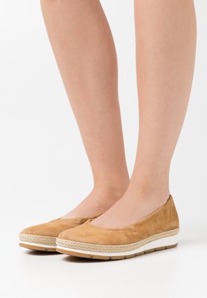 Piernas con zapatos planos de ante color beige, sin cordones, con detalles tejidos y suelas blancas sobre un fondo blanco.