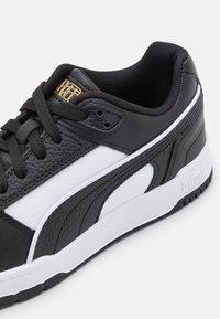 Chaussure de sport noire et blanche avec une tige en cuir texturé, une fermeture à lacets, et une semelle intermédiaire blanche avec des accents noirs. Détail du logo en or.