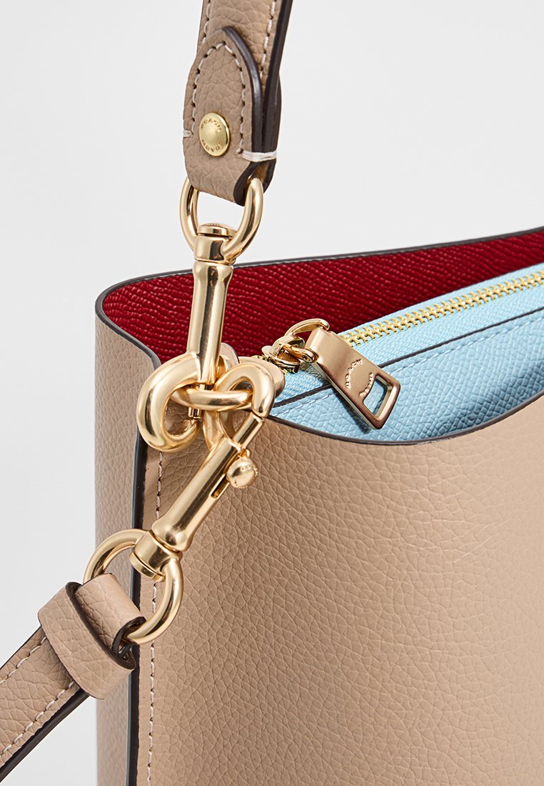 Sac en cuir tan avec un fermoir en métal doré, doublure intérieure rouge et un zip bleu ciel en haut. Texture lisse avec des accents cousus.