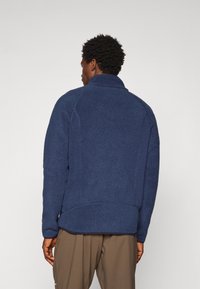 Navy blue fleece jacket with a textured surface, high collar, and a relaxed fit. Features a cinched waistband and subtle stitching details.