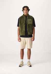 Green fleece vest with a high collar, zip front, and chest pocket. Worn over a black t-shirt, paired with beige shorts and sneakers.