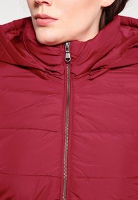 Burgundy puffer jacket with a zip closure, hood, and quilted texture. Features metallic zipper and stitching details on the surface.