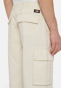 Beige cargobroek met meerdere zijzakken, gestructureerde stof en gebogen naaddetails. Beschikt over een logopatch op de achterzijde van de tailleband.