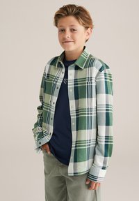 Zelena in bela karirana flanelna srajca z gumbom, nošena čez modro srajco; kombinirana z svetlimi hlačami za večplastni videz.