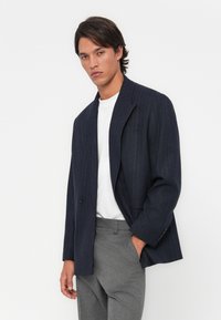 Navy pinstriped blazer in a tailored cut with a notched lapel, single button closure, and grey fitted trousers. Smooth fabric texture.