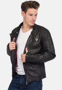 Zwarte leren motorjack met geborduurde schouder- en zijdetails, zilveren ritsen en een rode binnenvoering, gedragen over een wit t-shirt.